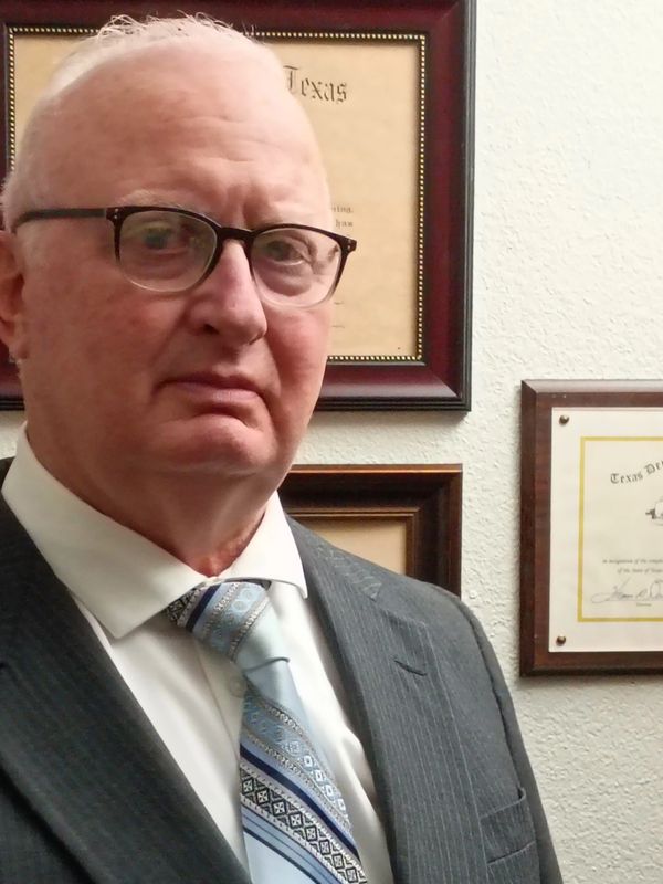 Older man in suit and glasses stands in front of framed certificates.