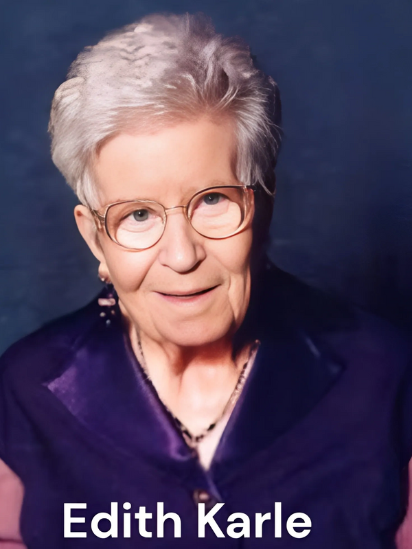 Portrait of an elderly woman with glasses wearing a purple outfit, smiling gently.