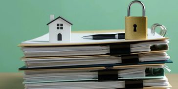 Stack of documents with a miniature house, pen, and padlock on top.
