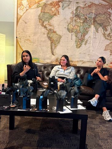 Three women sit on a couch with gift bags on a table in front of them.