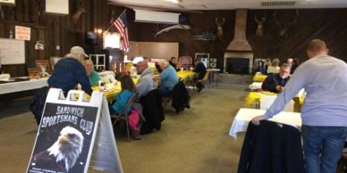 People gathered inside a hall with tables and a Sportsmans Club sign.