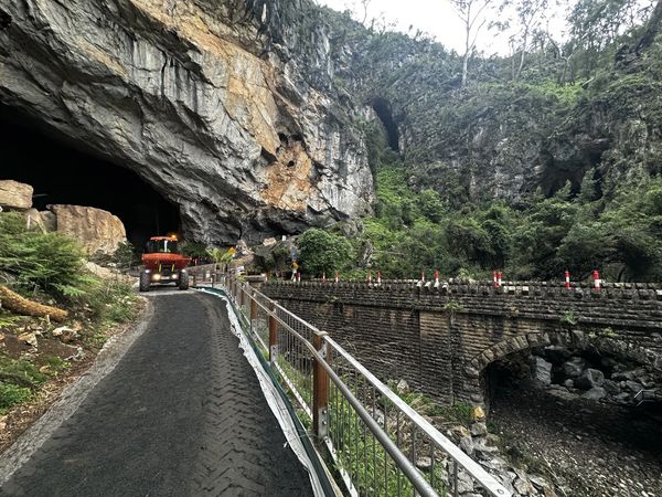 A vehicle enters a tunnel carved into a rocky mountain with a bridge on the side.