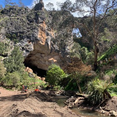 Large cave entrance surrounded by rocky cliffs and lush greenery on a sunny day.