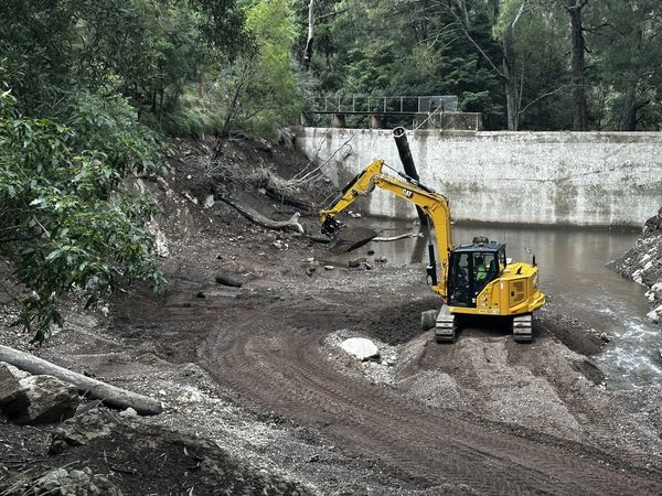 Yellow excavator working near a water dam in a forested area.