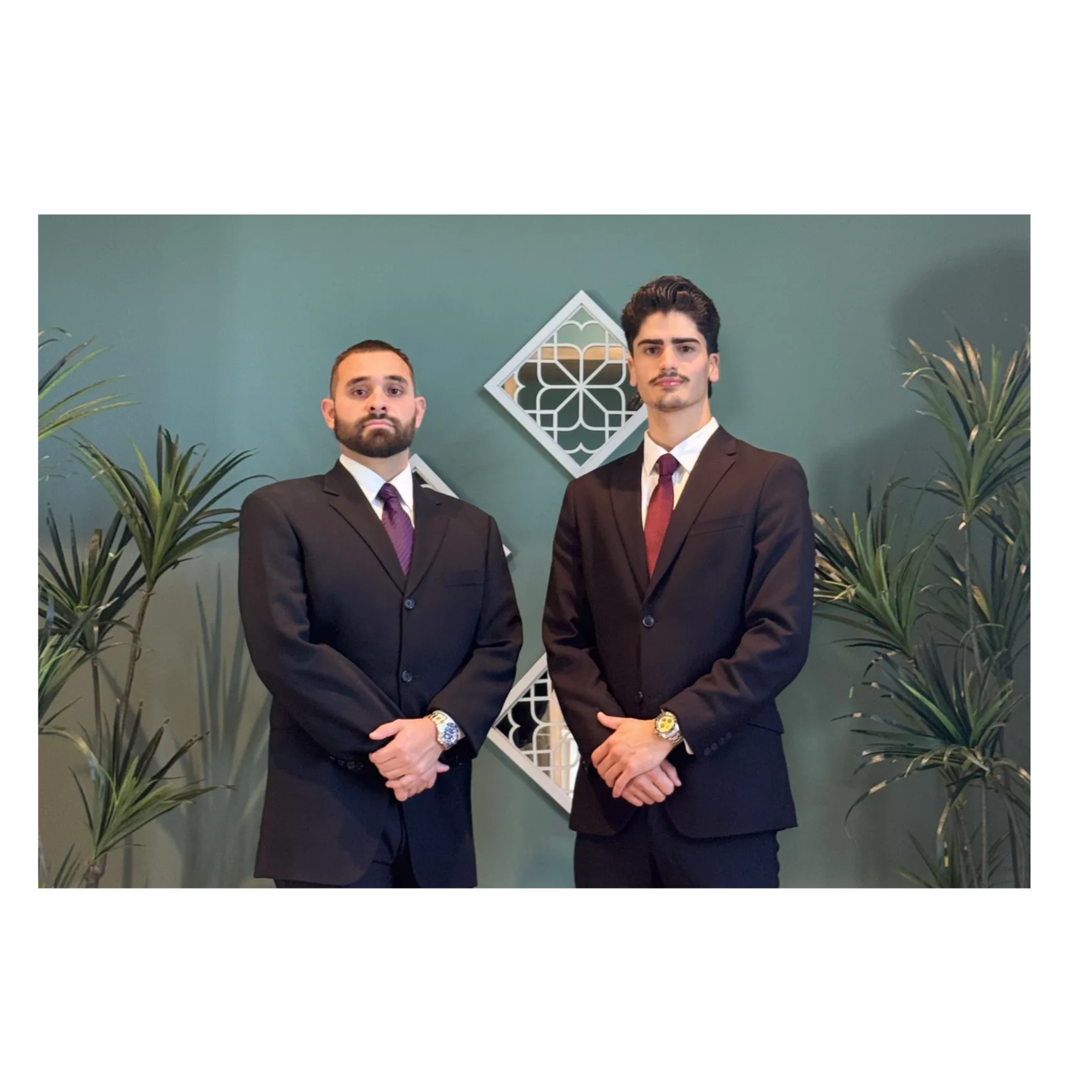 Two men in suits standing formally against a green wall with plants.