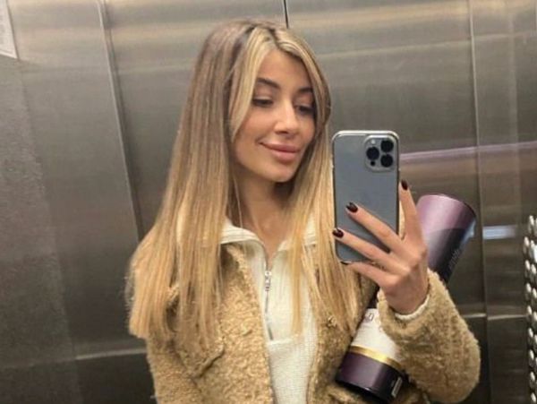 Young woman taking a mirror selfie in an elevator, wearing a beige jacket and black pants.
