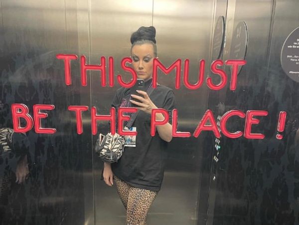 Person taking elevator selfie with neon text 'THIS MUST BE THE PLACE!'