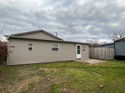 Backyard view of a small beige house with a concrete patio under a cloudy sky.