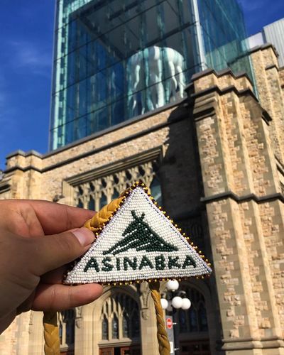 A hand holding up a beaded medallion that says "Asinabka".