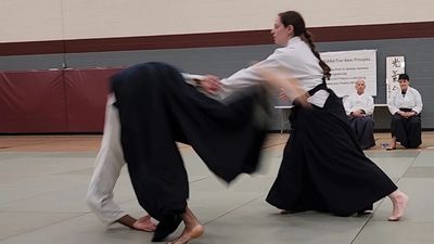 Sam-sensei, a short woman with a braid, is throwing a taller opponent, who is falling into a roll.