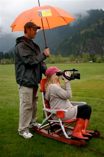 Woman sitting on chair in a field with video camera as she films a shot. Man holds umbrella over her