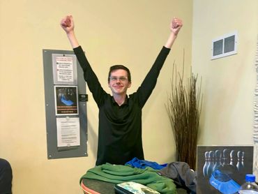 A man in black celebrates at a bowling alley with raised arms.