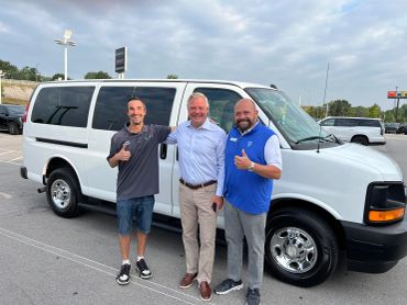 Three men smiling and giving thumbs up in front of a white van at a car dealership.