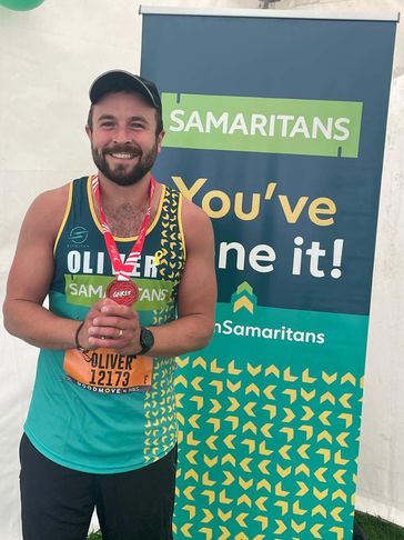 Smiling man named Oliver holds a medal in front of a Samaritans banner.