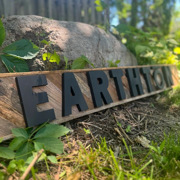 Reclaimed barnwood art sign spelling “EARTHTŌN” in bold black letters outdoors rested against a rock