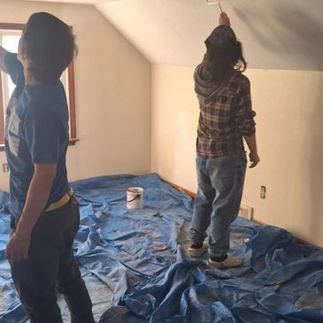Two people painting a ceiling with white paint in a room covered with blue drop cloths.