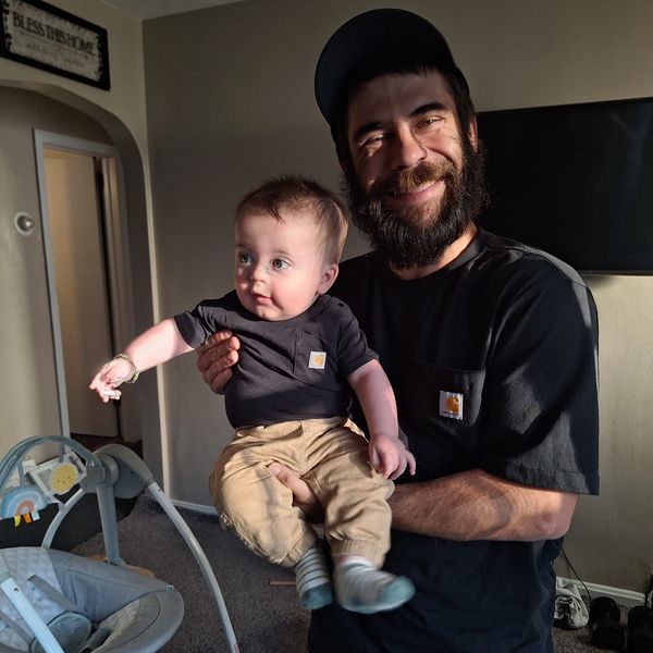 A bearded man happily holding a baby, both dressed in matching Carhartt outfits.