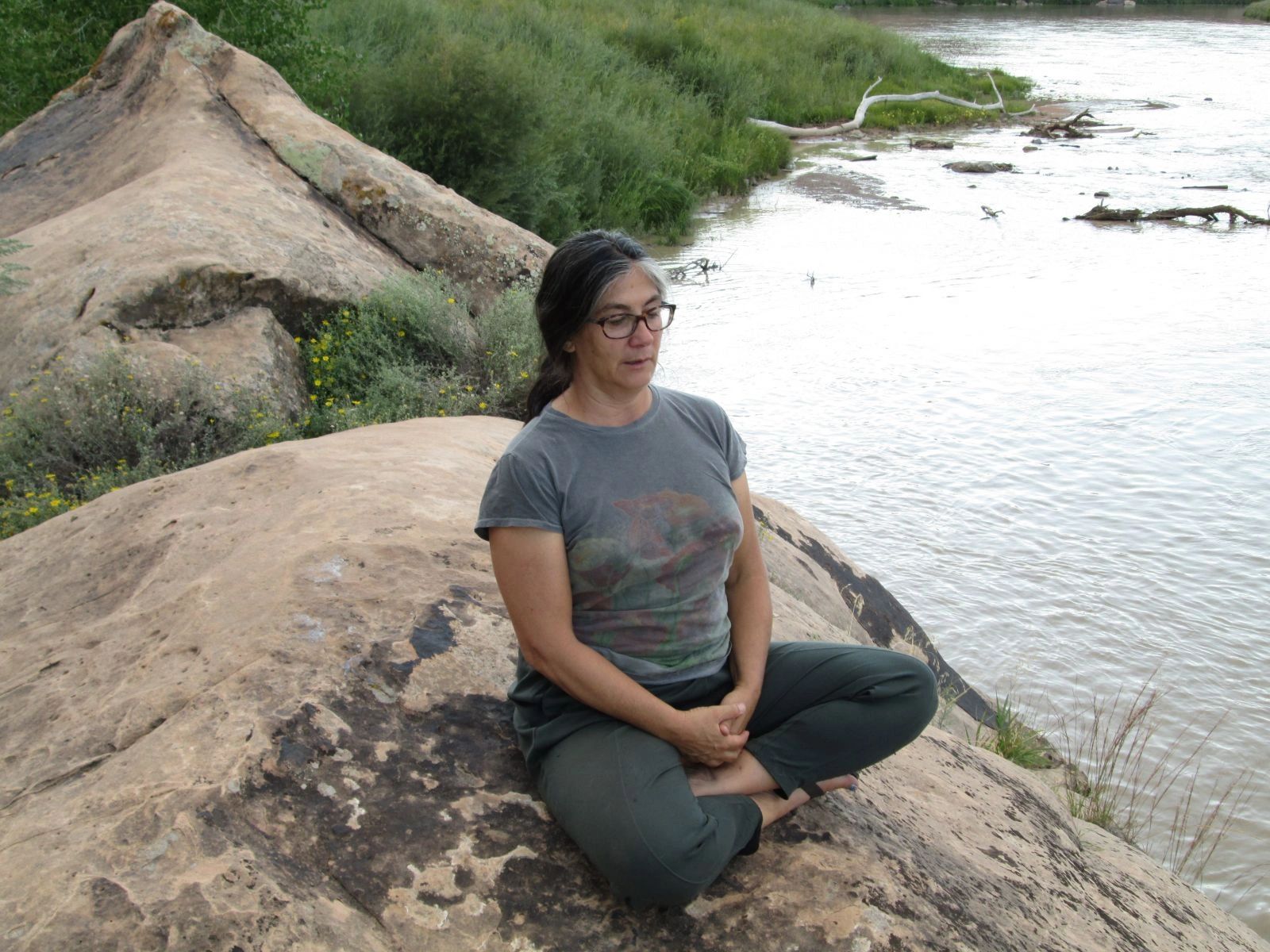Sue Schmidt Hypnotherapist sits on a large rock by a river.