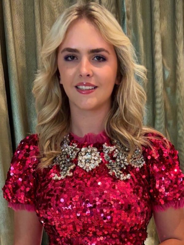 Young woman with blonde hair stands outdoors at sunset, wearing a white shirt.