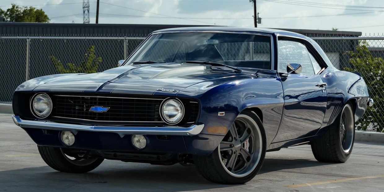 Classic blue Chevrolet Camaro parked on concrete with chain-link fence background.