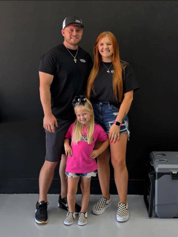 A smiling family of three posing against a black wall, dressed casually.