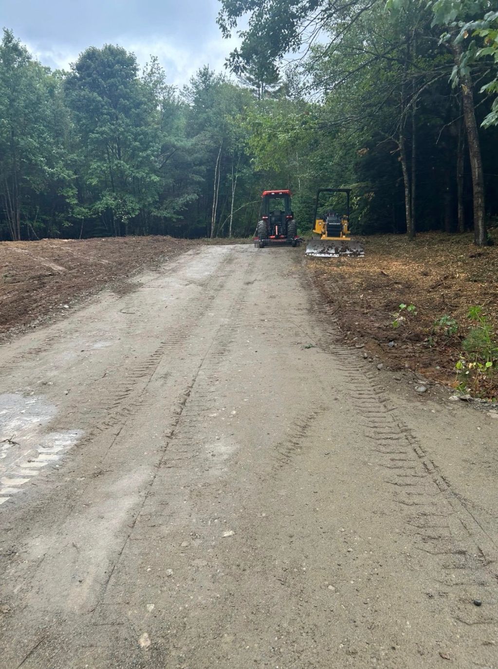 Cleared dirt road with two construction vehicles in a forest clearing.