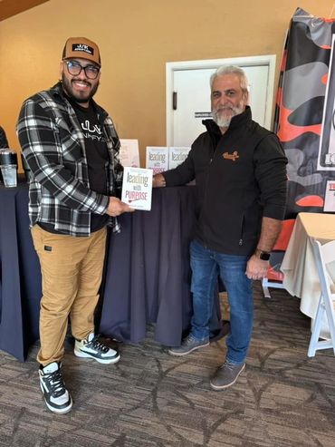Two men smiling, one holding a book titled 'Leading with Purpose.'