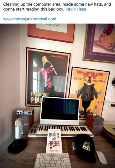 A tidy computer desk with a keyboard, microphone, book, and branded hats beneath vintage posters.