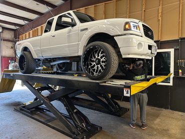 White pickup truck on a hydraulic lift with a mechanic working underneath.