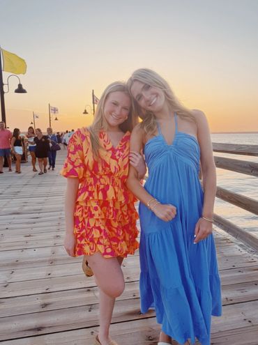 Two young women smiling on a wooden pier at sunset.