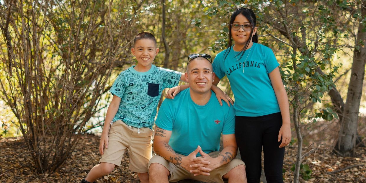 A father and his two kids sitting and standing next to each other in a nature during the day.
