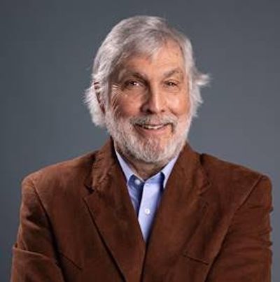 Smiling elderly man with gray hair and beard wearing brown jacket.