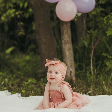 Smiling baby girl in a pink dress sitting on a blanket outdoors.