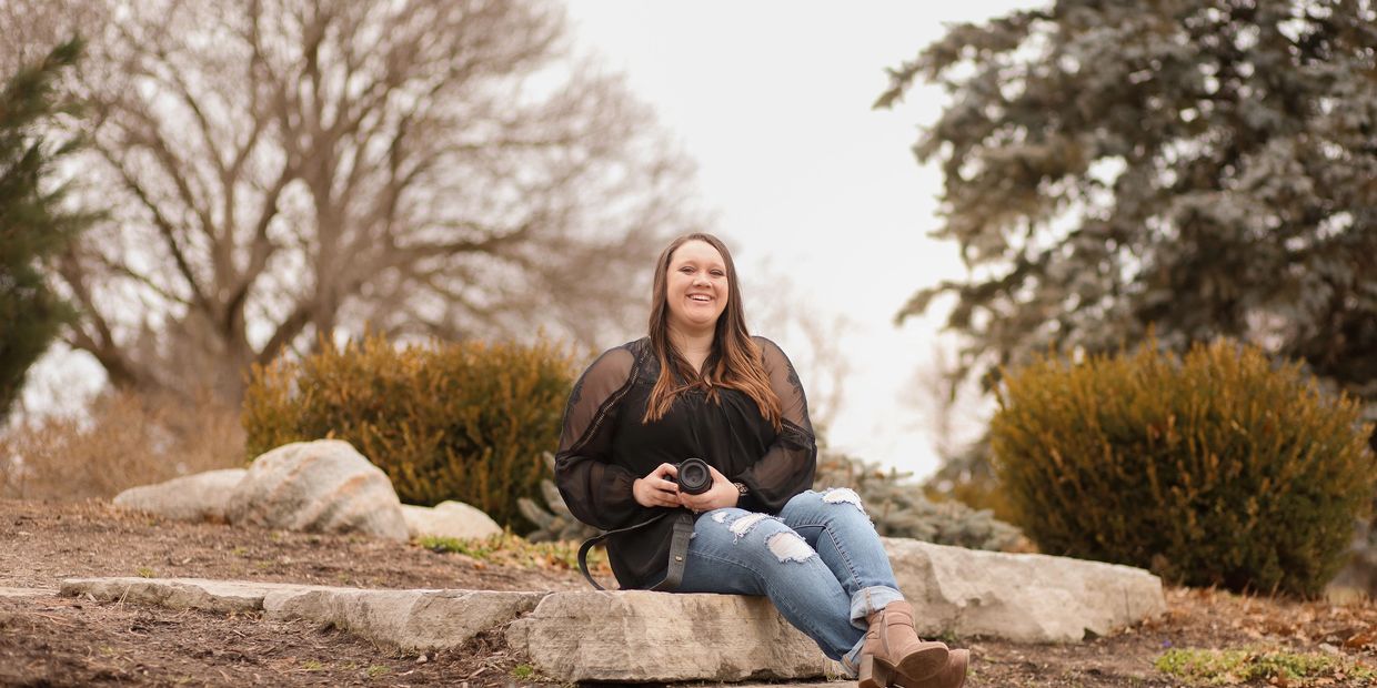 Smiling woman in a black top and ripped jeans sits on stone steps holding a camera.
