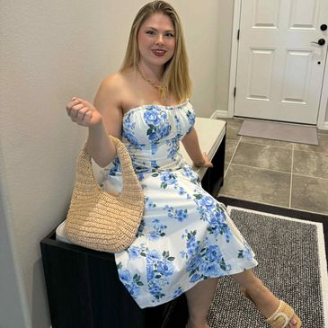 Woman in a white floral dress sitting indoors with a woven handbag.