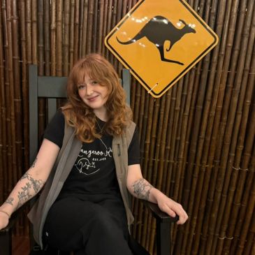 Young woman sits smiling in a chair with a kangaroo sign behind her.