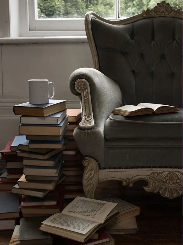 Cozy reading nook with a vintage armchair, stacks of books, and a coffee mug by a window with greenery outside.