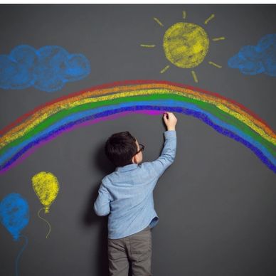 Child drawing a colorful rainbow, sun, and clouds with chalk on a blackboard.