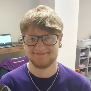 Young man with glasses and piercings smiling indoors.