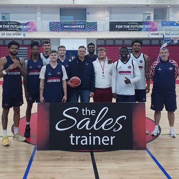 Basketball team posing with coaches behind a 'the Sales trainer' sign on court.
