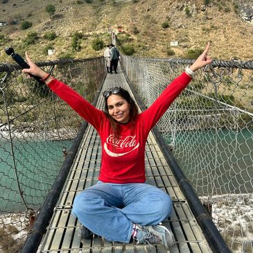 A woman in a red Coca-Cola sweatshirt sits cross-legged on a suspension bridge, smiling with arms raised.