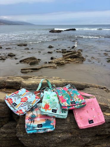 Colorful tropical-themed bags on a beach rock with ocean and seagull in background.