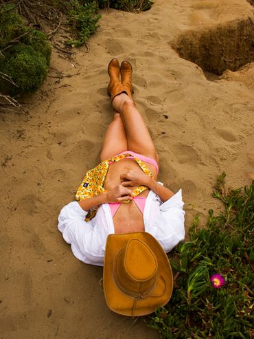 Person in a hat and boots relaxing on sandy ground near greenery.