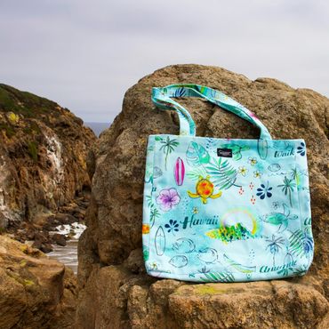 Colorful Hawaii-themed tote bag resting on rocky coastal cliffs.