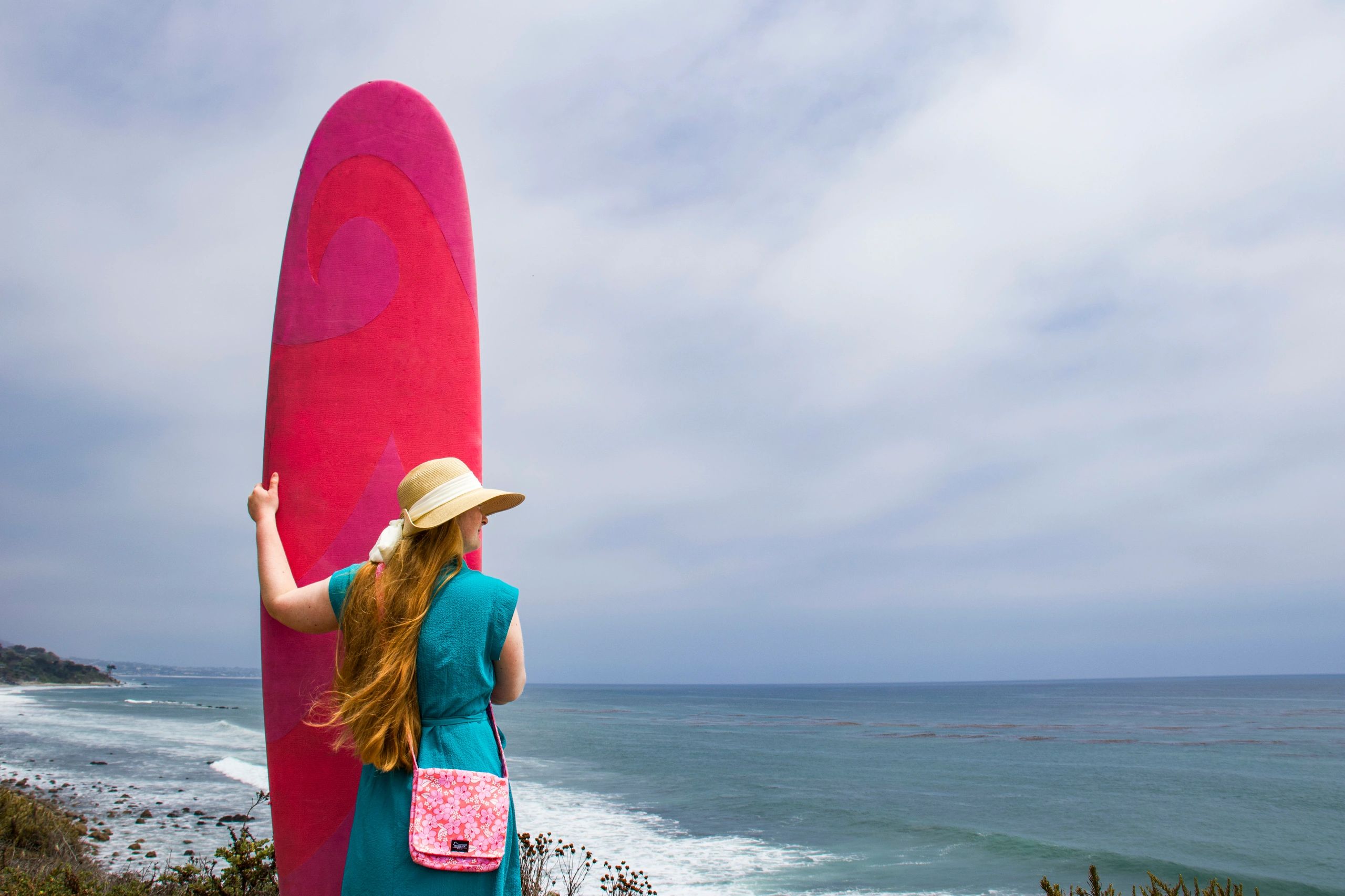 Woman in turquoise dress holding pink surfboard by the ocean under cloudy sky.