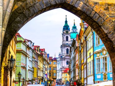 View of colorful historic buildings and a church tower framed by an old stone arch.