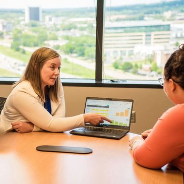 Two women discussing data charts on a laptop in an office with a city view.