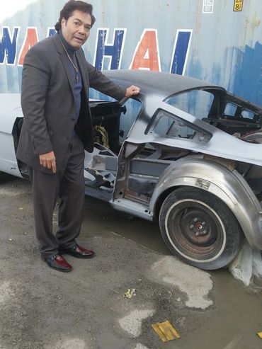 A man in a suit stands next to a stripped-down car frame outdoors.