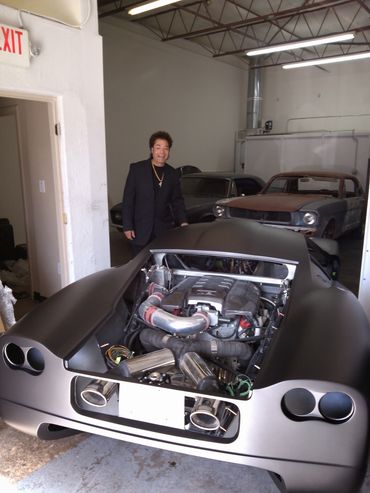 Man in black suit standing next to a custom car with exposed engine in a garage.