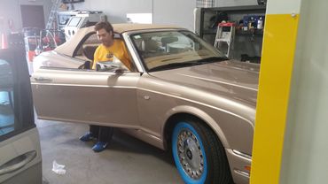 Man in yellow shirt getting into a beige convertible car with blue tire rims indoors.